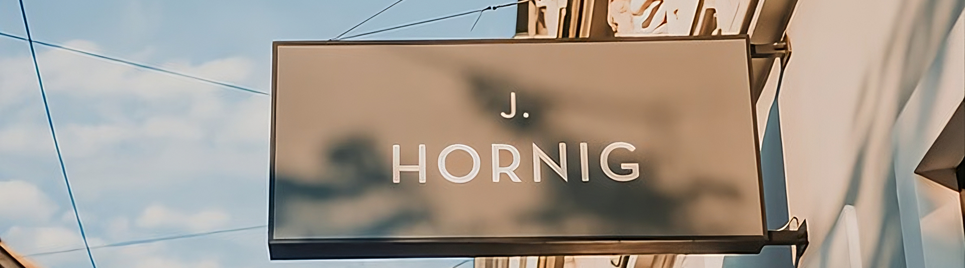 Schild für J. Hornig, das vor einem stilvollen Gebäude vor blauem Himmel hängt und Branding und städtische Architektur zeigt.