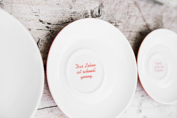 Weiße Keramikteller mit roter Beschriftung, die nachdenkliche Zitate auf einem rustikalen Holzhintergrund zeigen.