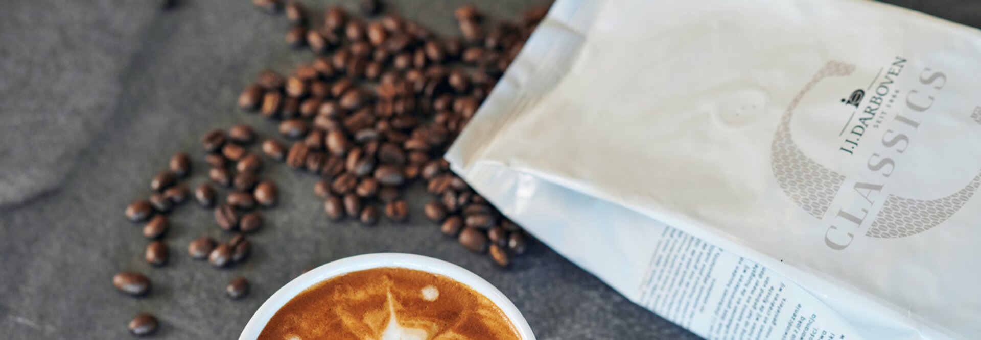 Latte so zložitým umeleckým dizajnom, obklopené kávovými zrnami a vreckom kávy J.J. Darboven Classics.