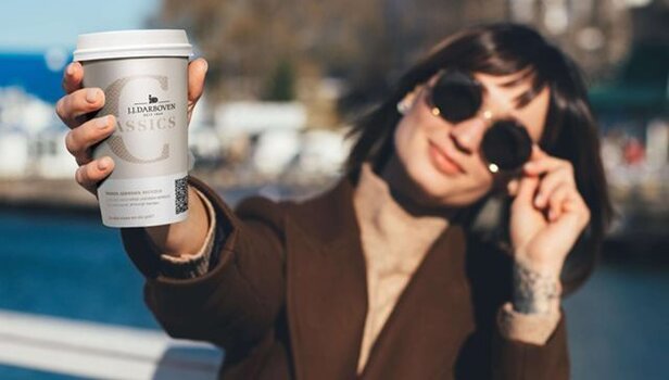Eine Frau genießt einen Kaffee zum Mitnehmen mit einer stilvollen Sonnenbrille am Wasser, die das JJD Classics-Branding auf der Tasse zeigt.