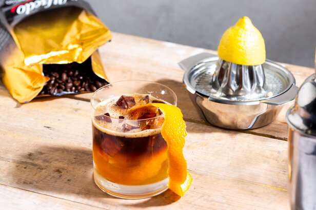 Espresso-Amaretto-Sour-Cocktail mit Orangenspirale, Kaffeebohnen und einer Zitronenpresse auf einem rustikalen Holztisch.