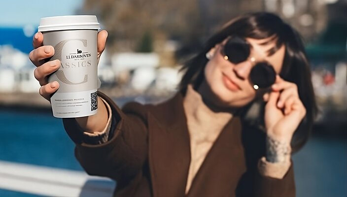 A woman enjoys a takeaway coffee while wearing sunglasses, set against a picturesque outdoor background.