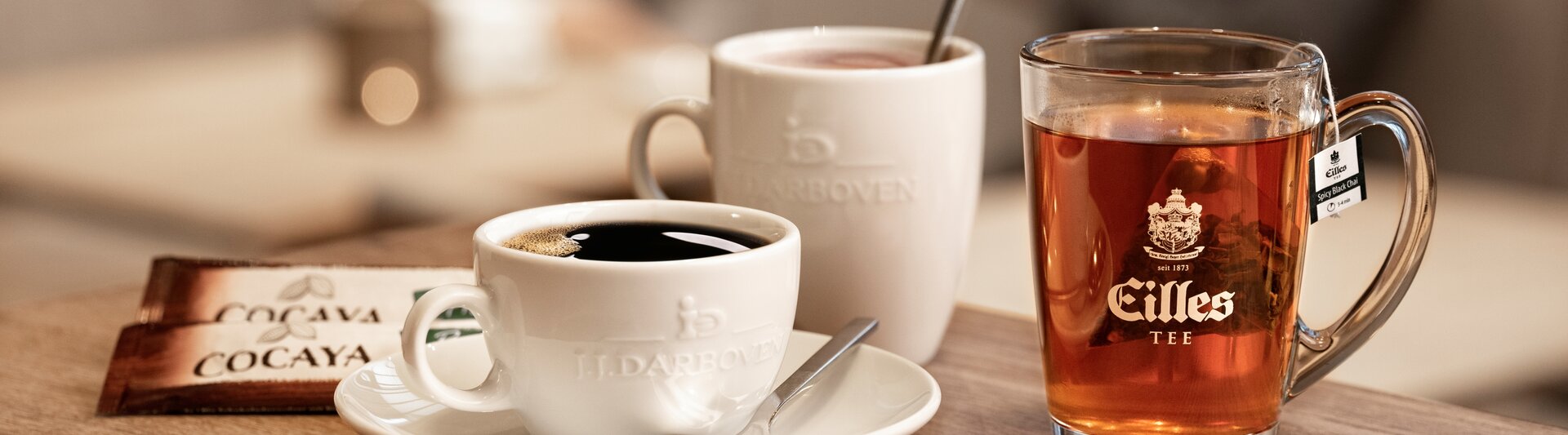 Ein schwarzer Kaffee und ein Glas Tee stehen auf einem Holztisch, und im verschwommenen Hintergrund unterhalten sich zwei Personen.