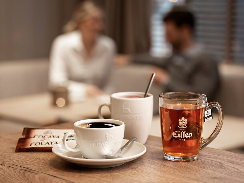 A black coffee and a glass of tea sit on a wooden table, with two people chatting in the blurred background.