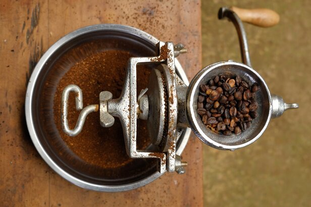 Vintage-Kaffeemühle mit gerösteten Bohnen, bereit zum Mahlen auf einem Holztisch, umgeben von frisch gemahlenem Kaffee.