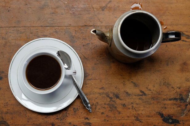 Kaffeetasse mit Untertasse und Löffel neben einer Teekanne auf einem rustikalen Holztisch, der einen warmen Rahmen für ein Getränk bietet.