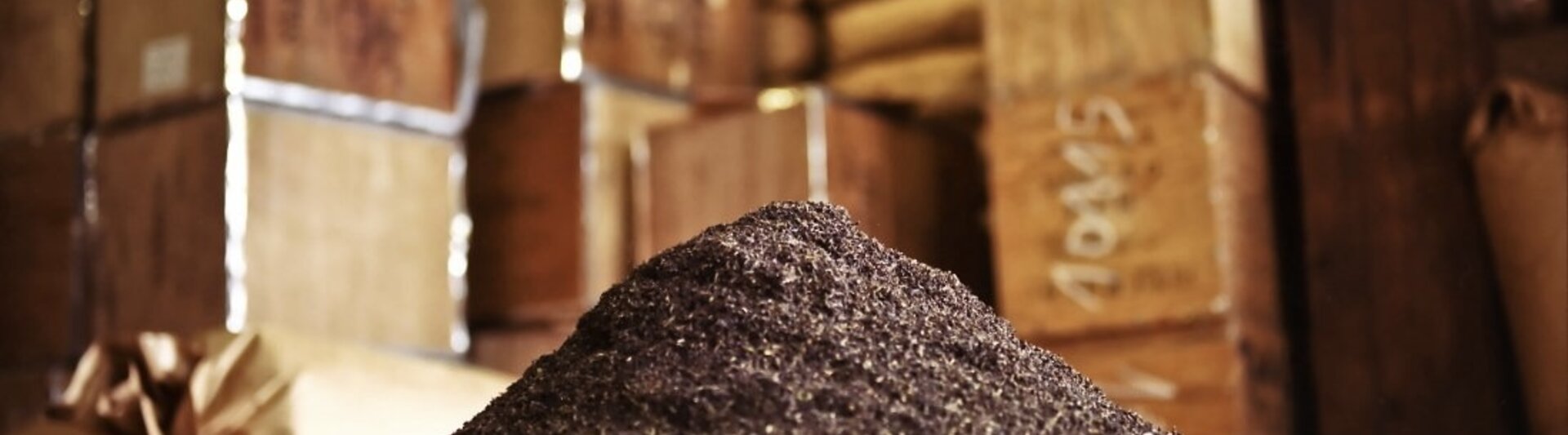 Mountain of dark ground spices in a rustic storage area with stacked boxes in the background.