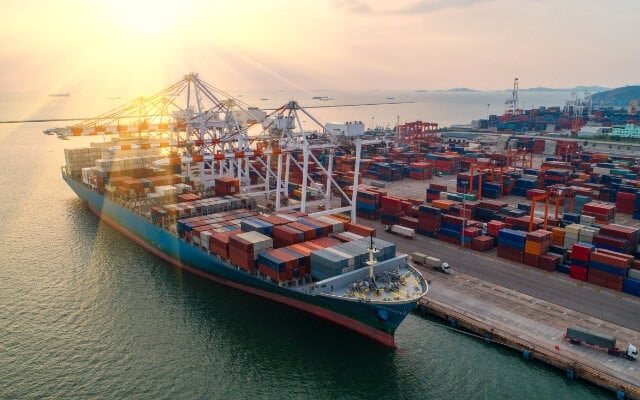 Ein Containerschiff, das an einem belebten Hafen angedockt hat, beladen mit Fracht unter einem leuchtenden Sonnenuntergangshimmel.