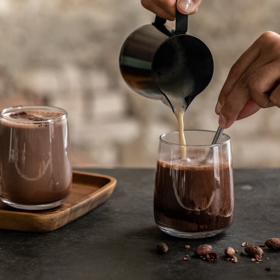 Verser un chocolat chaud mousseux dans un verre, avec des fèves de cacao et une truffe au chocolat sur la table.