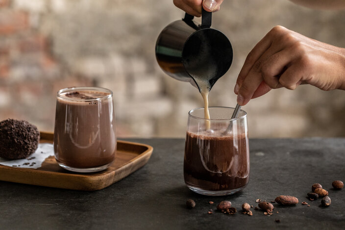 Pouring frothy hot chocolate into a glass, with cocoa beans and a chocolate truffle on the table.