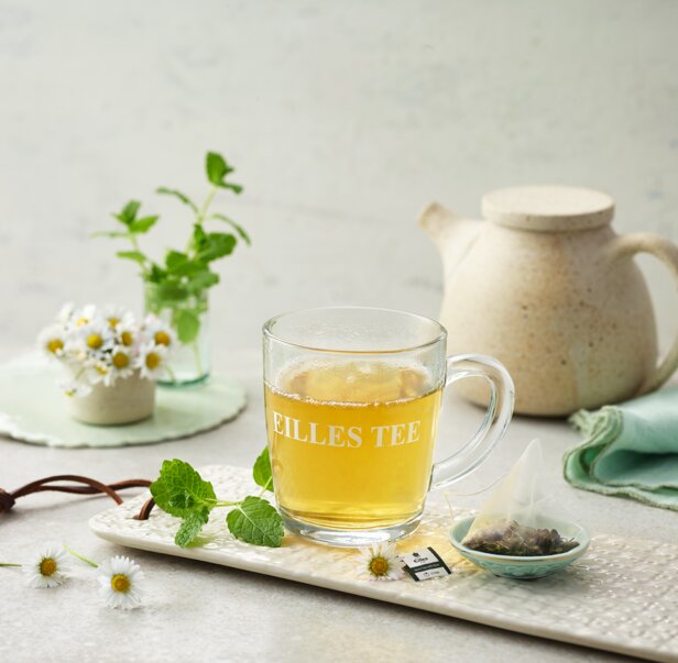 Verfrissende EILLES TEE in een glazen beker, vergezeld van muntblaadjes en madeliefjes, perfect om in de lente van te genieten.