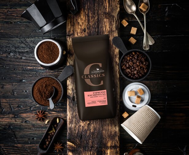 Balenie kávy Espresso Hausröstung obklopené kávovými zrnami, mletou kávou a kockami cukru na rustikálnom drevenom pozadí.