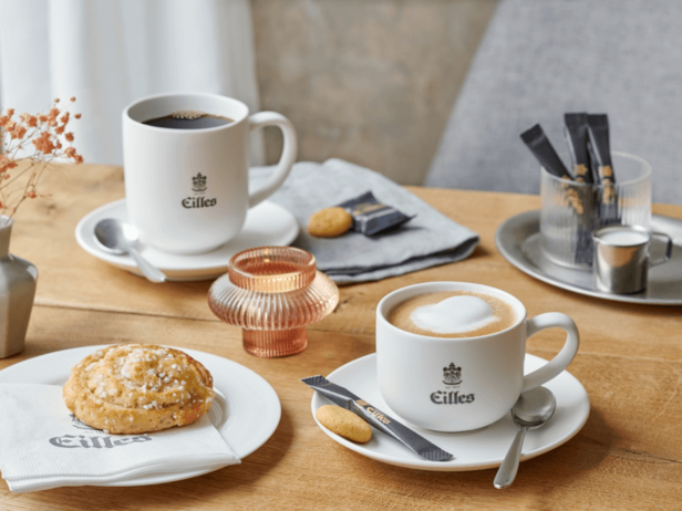 Elegantná káva a zákusky Eilles na drevenom stole, s kapučínom, čiernou kávou a rozkošným koláčikom.