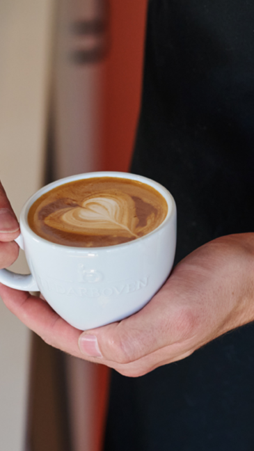 Barista holding a latte with heart-shaped art in a white cup, showcasing expert coffee craftsmanship.
