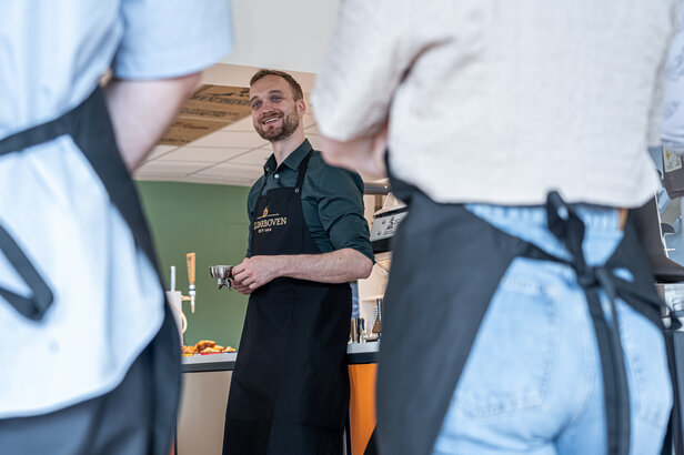 Barista serving coffee in a café, engaging with customers in aprons, showcasing a friendly atmosphere.