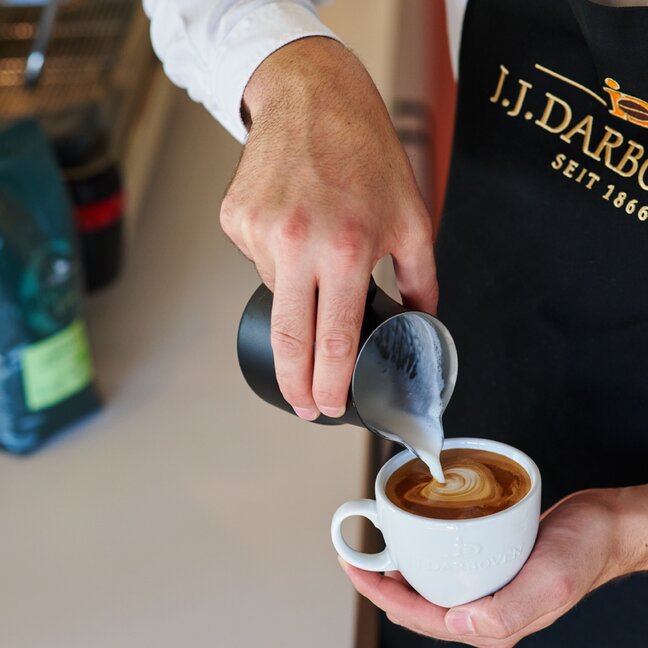 Barista pouring microfoam into a coffee cup, creating beautiful latte art with J.J. Darboven coffee beans.
