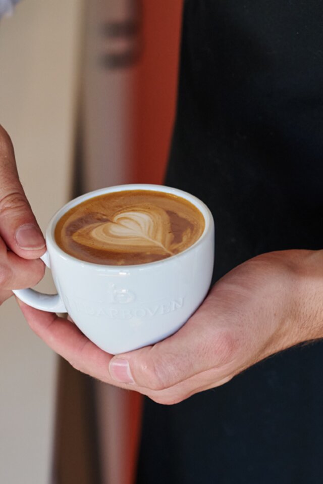 Barista předvádí latte art v bílém šálku s motivem srdce na krémové kávě. Viditelná značka J.J. Darboven.