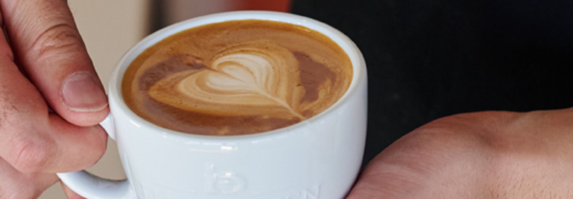 Barista předvádí latte art v bílém šálku s motivem srdce na krémové kávě. Viditelná značka J.J. Darboven.