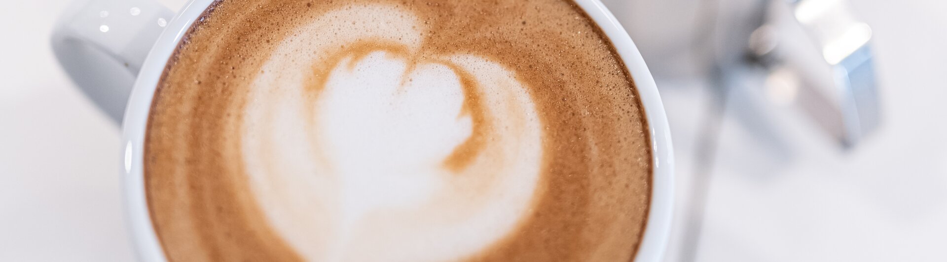A beautiful latte with a heart design, accompanied by metal milk frothing jugs on a clean white surface.