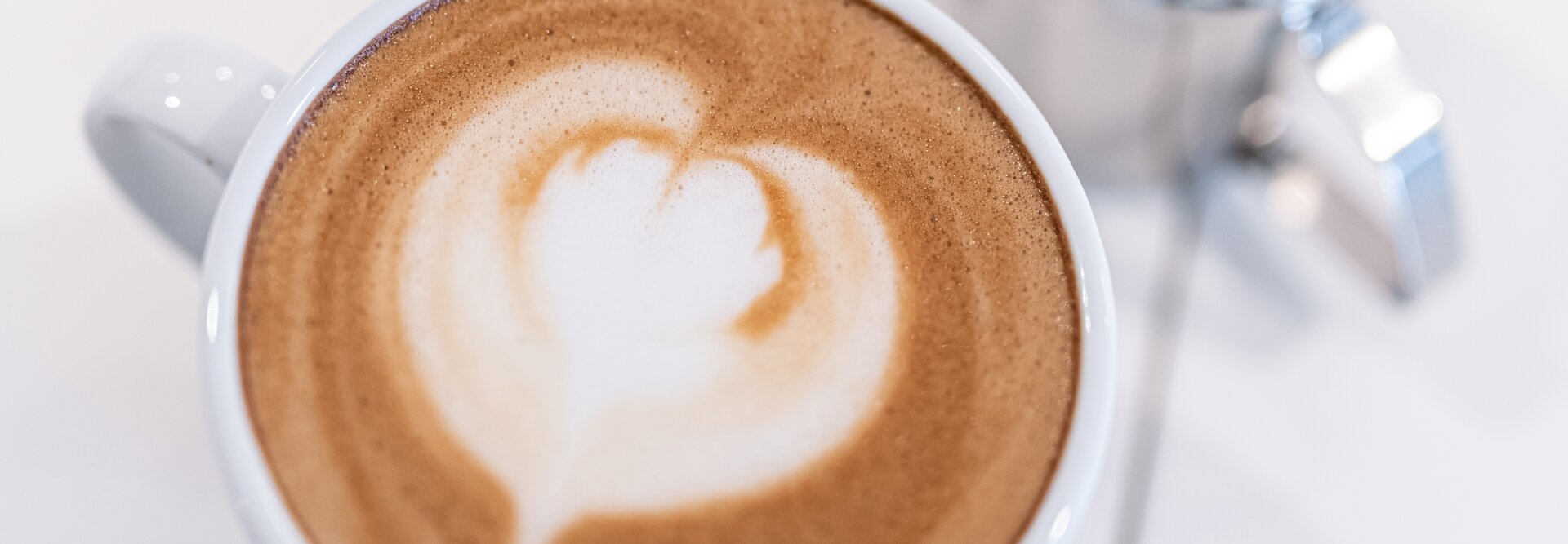 Ein schöner Milchkaffee mit Herzmotiv, begleitet von Metallkännchen zum Aufschäumen von Milch auf einer sauberen weißen Oberfläche.
