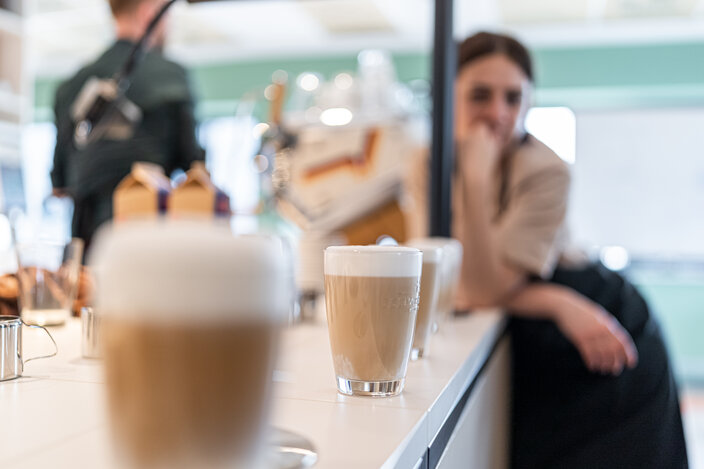 Barista serving coffee in a modern café, with lattes lined up on the counter and a customer in the background.