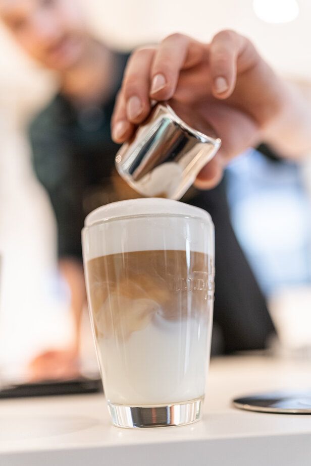 Ein Barista gießt Milch über ein geschichtetes Kaffeegetränk in einem Glas und zeigt die Technik der Latte Art in einer Caféumgebung.