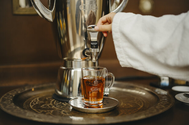 Ruka nalieva čaj zo štýlového dávkovača do skleneného hrnčeka a predvádza teplý nápoj na ozdobnom podnose.