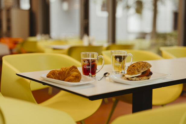 Frühstückstisch mit einem Croissant, einem Sandwich und zwei Tassen Tee in einem modernen Café mit gelben Stühlen.