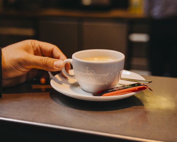 Une main tient une tasse de café blanc sur le comptoir d'un café, accompagnée d'une petite cuillère et d'un bâton de sucre.
