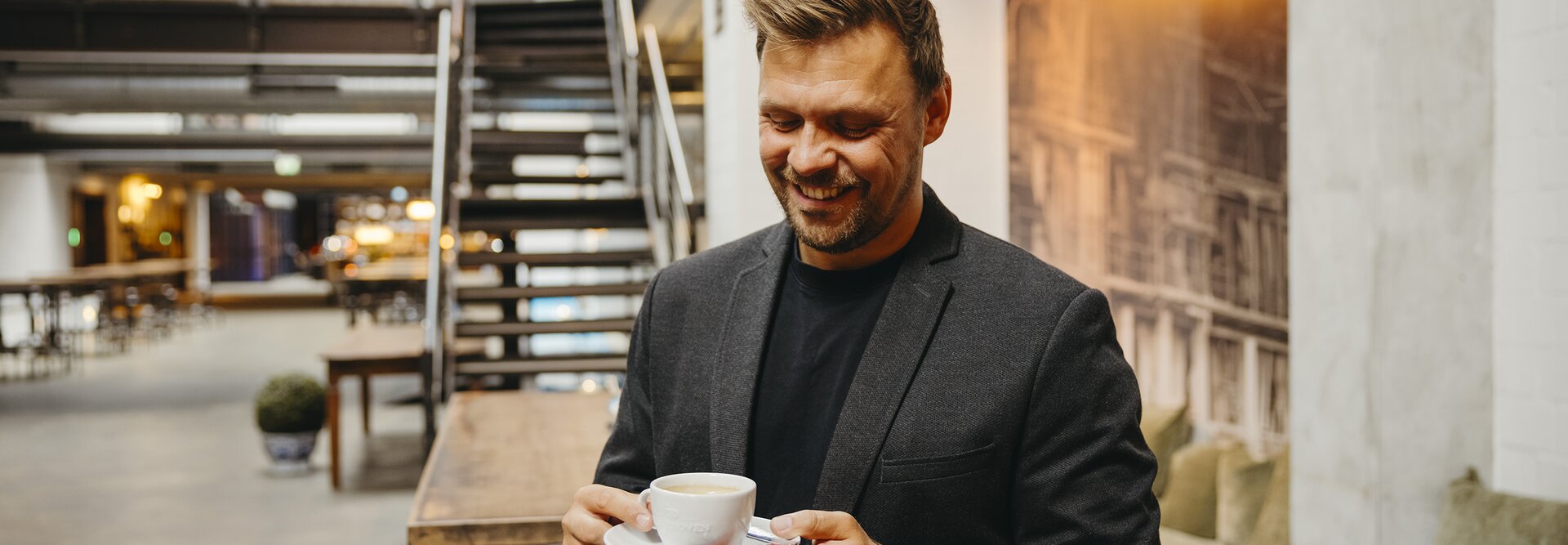 Ein lächelnder Mann in einem Blazer genießt eine Tasse Kaffee in einem stilvollen, modernen Café mit Holzmöbeln.