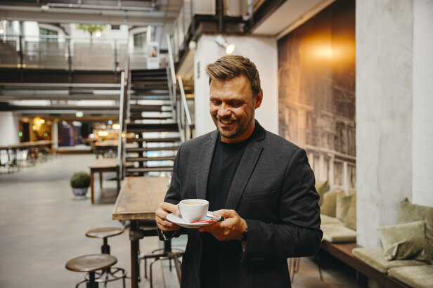 Een glimlachende man in een blazer geniet van een kopje koffie in een stijlvol, modern café met houten meubilair.