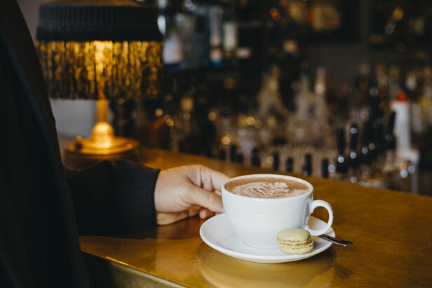 Stilvolle Kaffeetasse mit Latte Art, begleitet von einem Macaron, vor einer warmen, einladenden Bar-Kulisse.