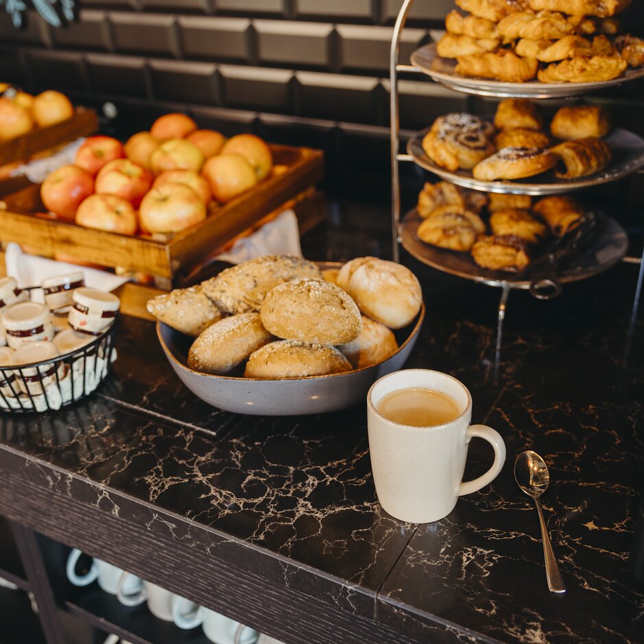 Eine Auswahl an frischem Gebäck, Brötchen und Äpfeln begleitet von einer Tasse Tee an einem stilvollen Cafétisch.