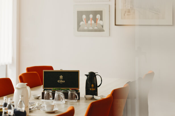 Eleganter Besprechungsraum mit orangefarbenen Stühlen, einem Teeservice und Gläsern auf einem Holztisch, der eine einladende Atmosphäre schafft.