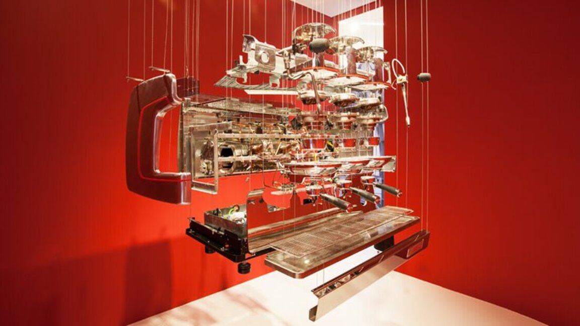 Artistic display of a disassembled espresso machine suspended against a vibrant red background, showcasing its intricate parts.