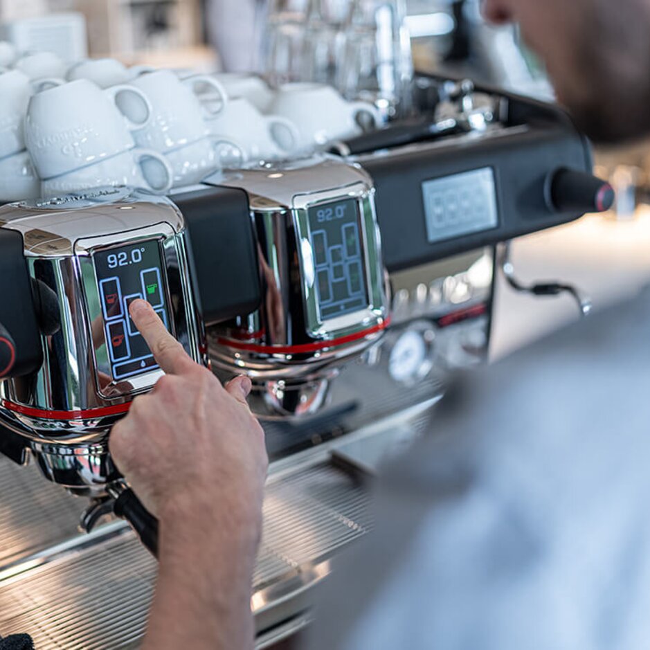 Barista adjusting temperature settings on a high-end espresso machine, with stacked cups in the background.