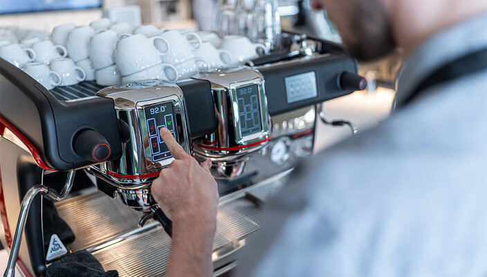 Barista bei der Temperatureinstellung an einer hochwertigen Espressomaschine, mit gestapelten Tassen im Hintergrund.