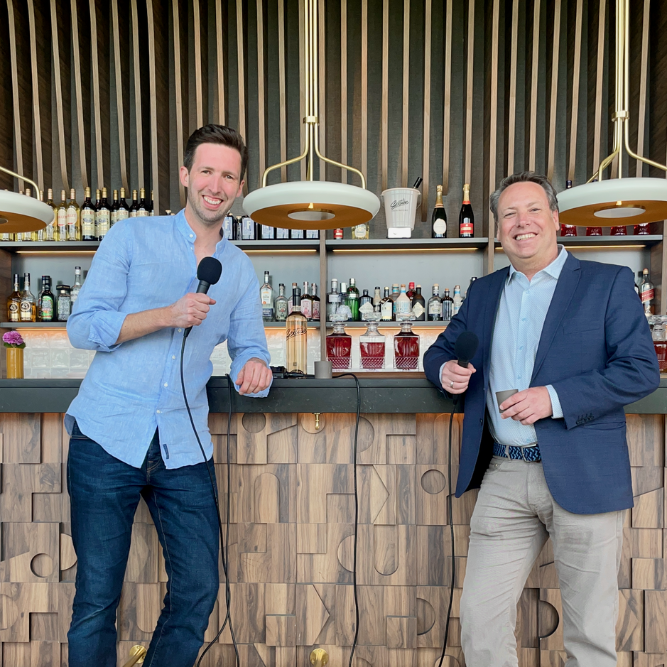 Two men holding microphones pose at a stylish bar, showcasing a vibrant selection of spirits behind them.