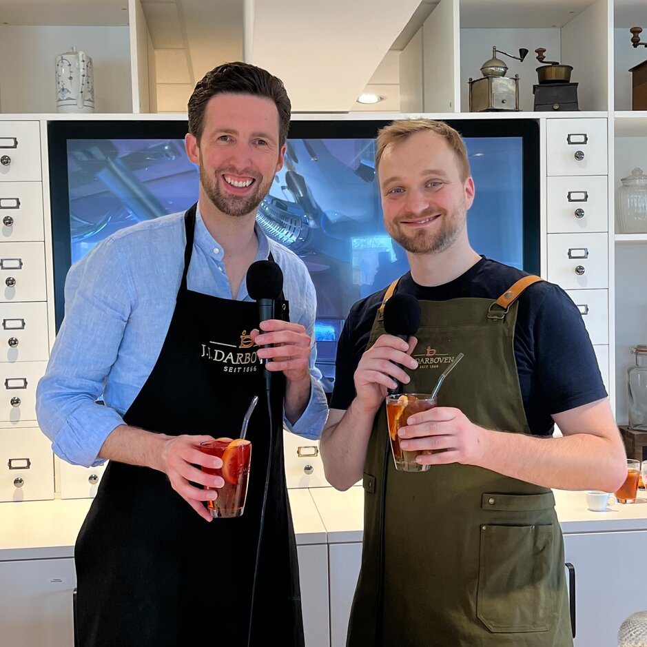 Zwei lächelnde Gastgeber in Schürzen halten Getränke und Mikrofone in der Hand, bereit, in einer lebhaften Podcast-Umgebung über Cocktails zu diskutieren.