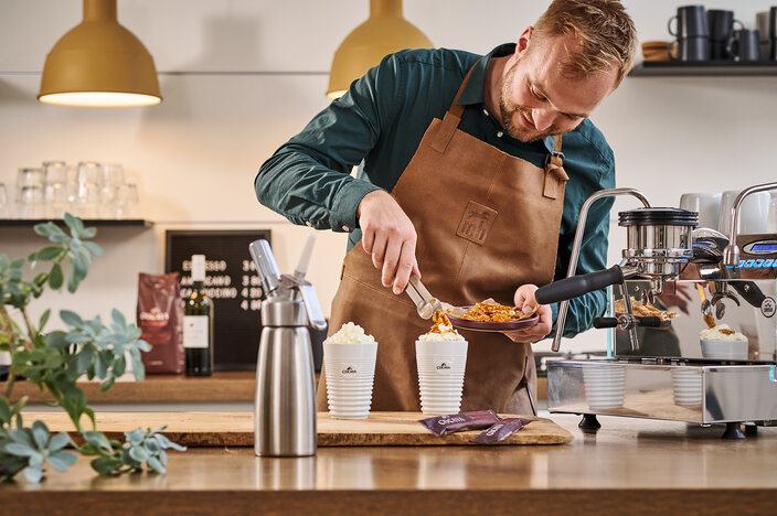 Barista in an apron garnishing desserts with toppings at a coffee bar, showcasing premium drinks and a stylish café setup.