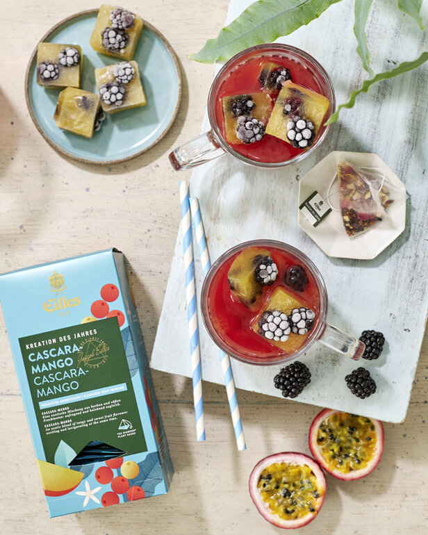 Erfrischender Cascara-Mango-Eistee mit Fruchteiswürfeln, Maracuja-Scheiben und Brombeeren, perfekt für den Sommer.