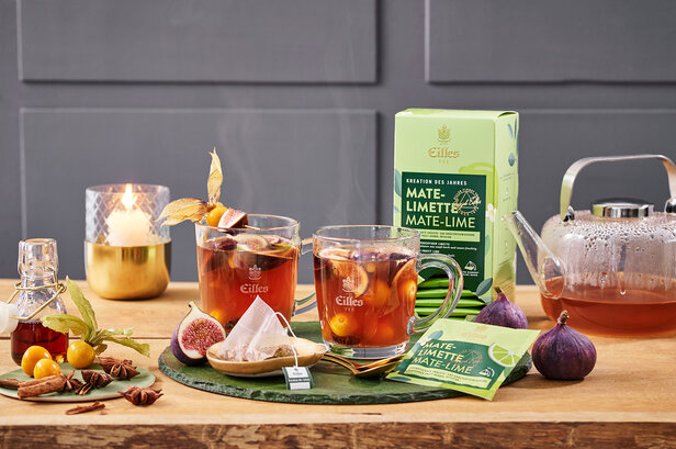 Warmer Mate-Limette-Tee mit Feigen und Gewürzen, mit der Verpackung von Gille und einem Kessel auf einem Holztisch. Perfekt für gemütliche Momente.