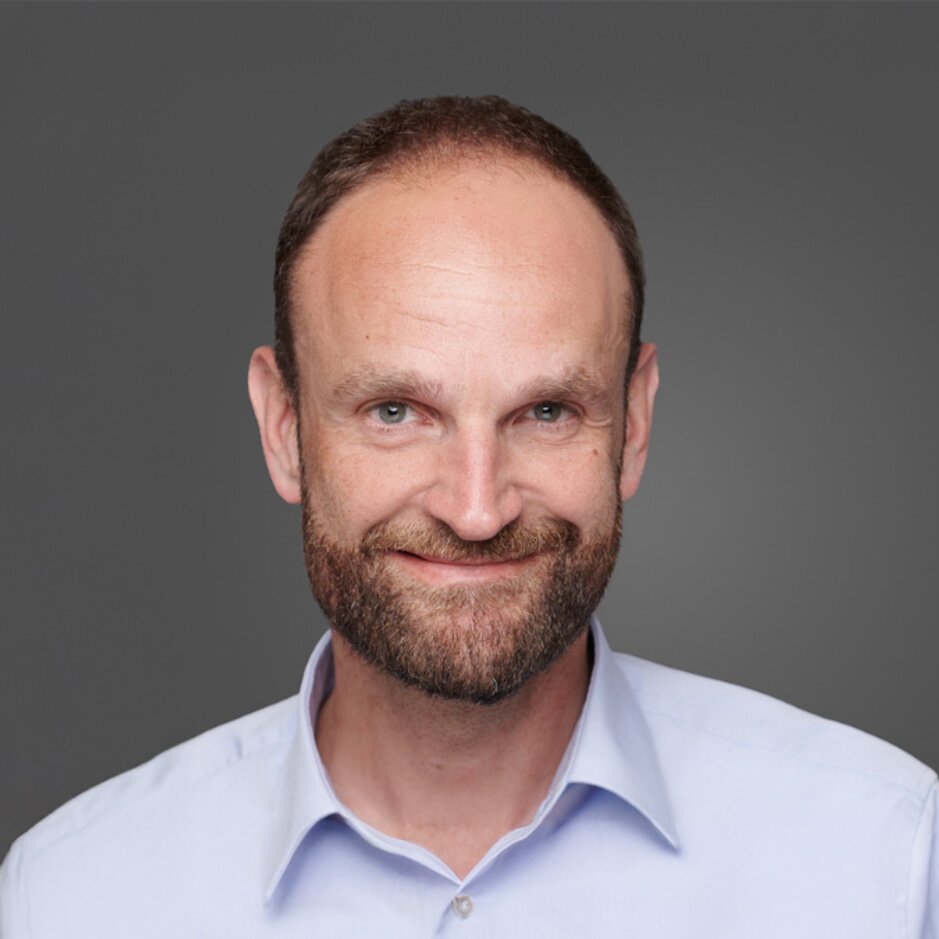 Smiling man with a beard wearing a light blue shirt, set against a grey background. Suitable for professional profiles.