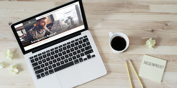 Ordinateur portable sur un bureau en bois affichant un site web de tarification du café, avec à côté une tasse de café et un bloc-notes portant l'inscription "TASSENPREIS ?