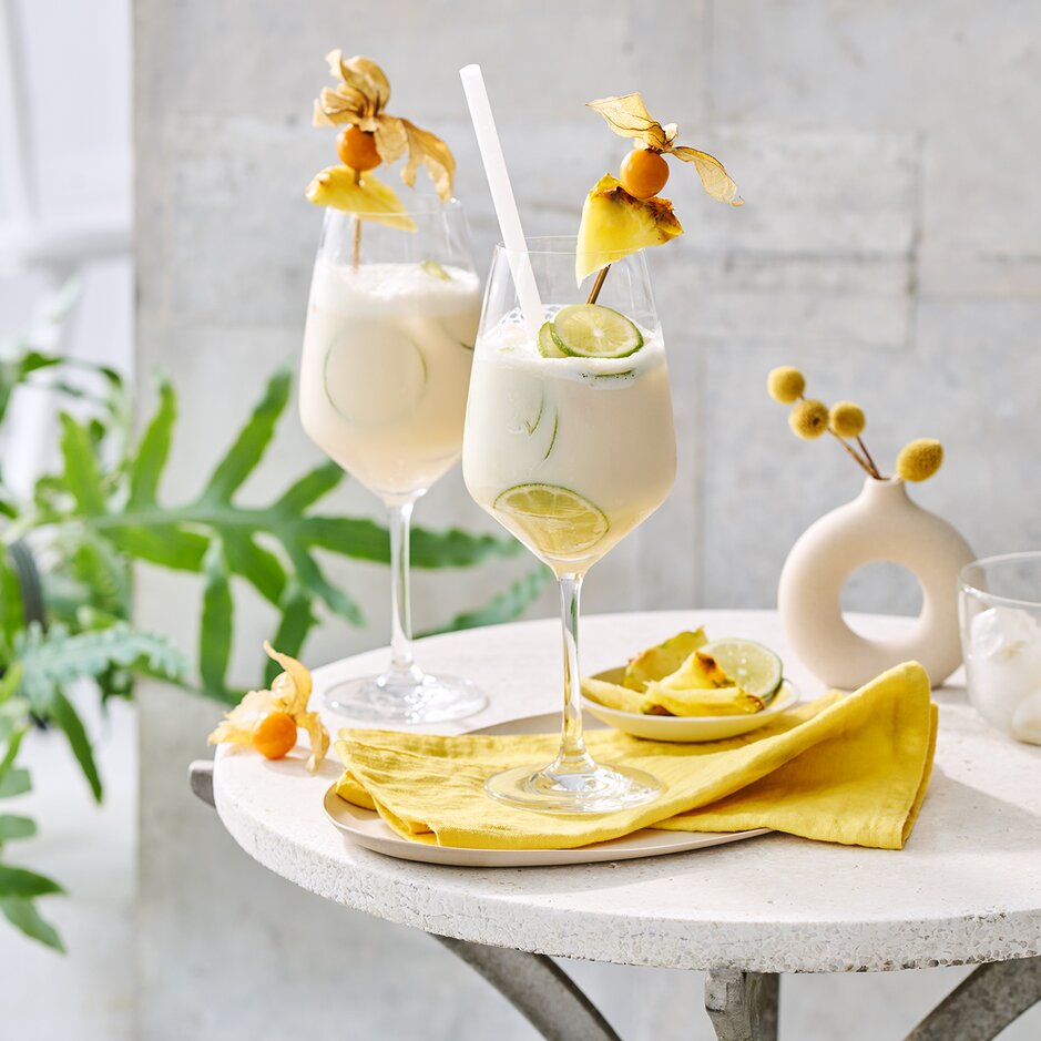 Zwei elegante Pina-Colada-Cocktails, garniert mit Ananas und Limetten, serviert auf einem stilvollen Tisch mit Grünzeug.