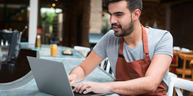 Ein junger Mann in einer Schürze arbeitet mit einem Laptop in einem modernen Restaurant und setzt damit auf die Digitalisierung im Gastgewerbe.