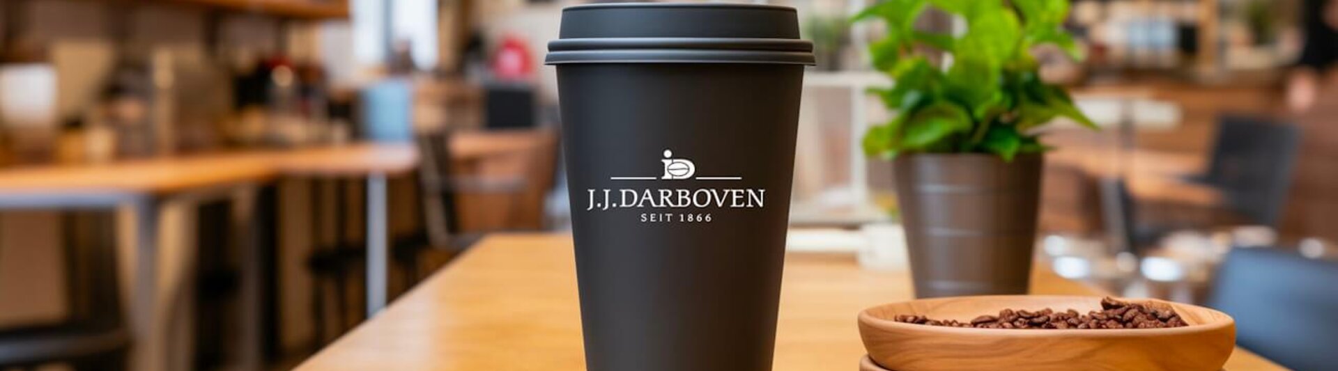 Wiederverwendbarer Kaffeebecher auf einem Holztisch, der das Branding von J.J. Darboven in einer stilvollen Caféumgebung zeigt.