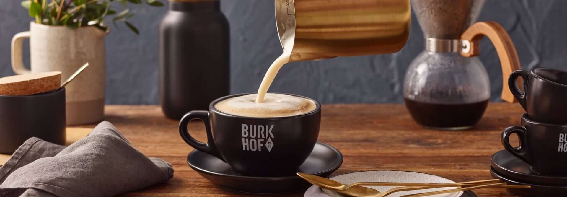 Samtige Milch in einen Cappuccino gießen, in stilvollem Café-Ambiente mit Burkhof-Accessoires.