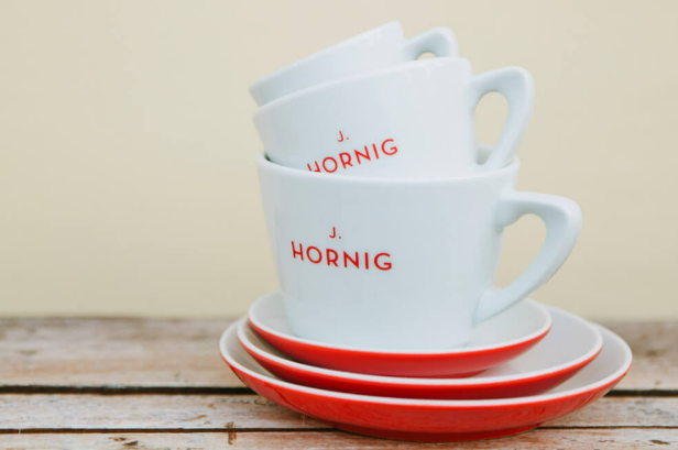 Stacked white coffee cups with red accents and saucers on a wooden table, featuring the name "J. Hornig".