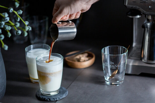 Pouring coffee into a glass of milk, surrounded by elegant glassware and a stylish coffee machine.